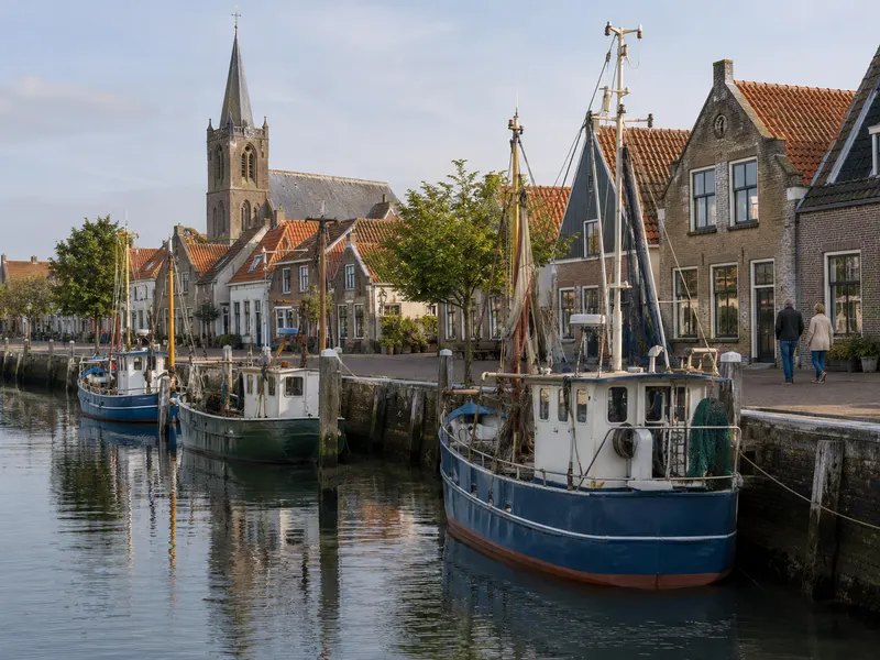 Een klein havenstadje: boven lage bakstenen huizen met rode daken staat een hoge kerktoren. In de haven liggen drie vissersboten, blauw en groen. Twee mensen lopen op de kade.